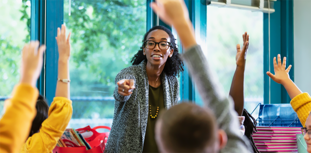 photo of dark-skinned middle-aged teacher with glasses and medium length curly hair standing at the front of a classroom and pointing to a student to call on them.